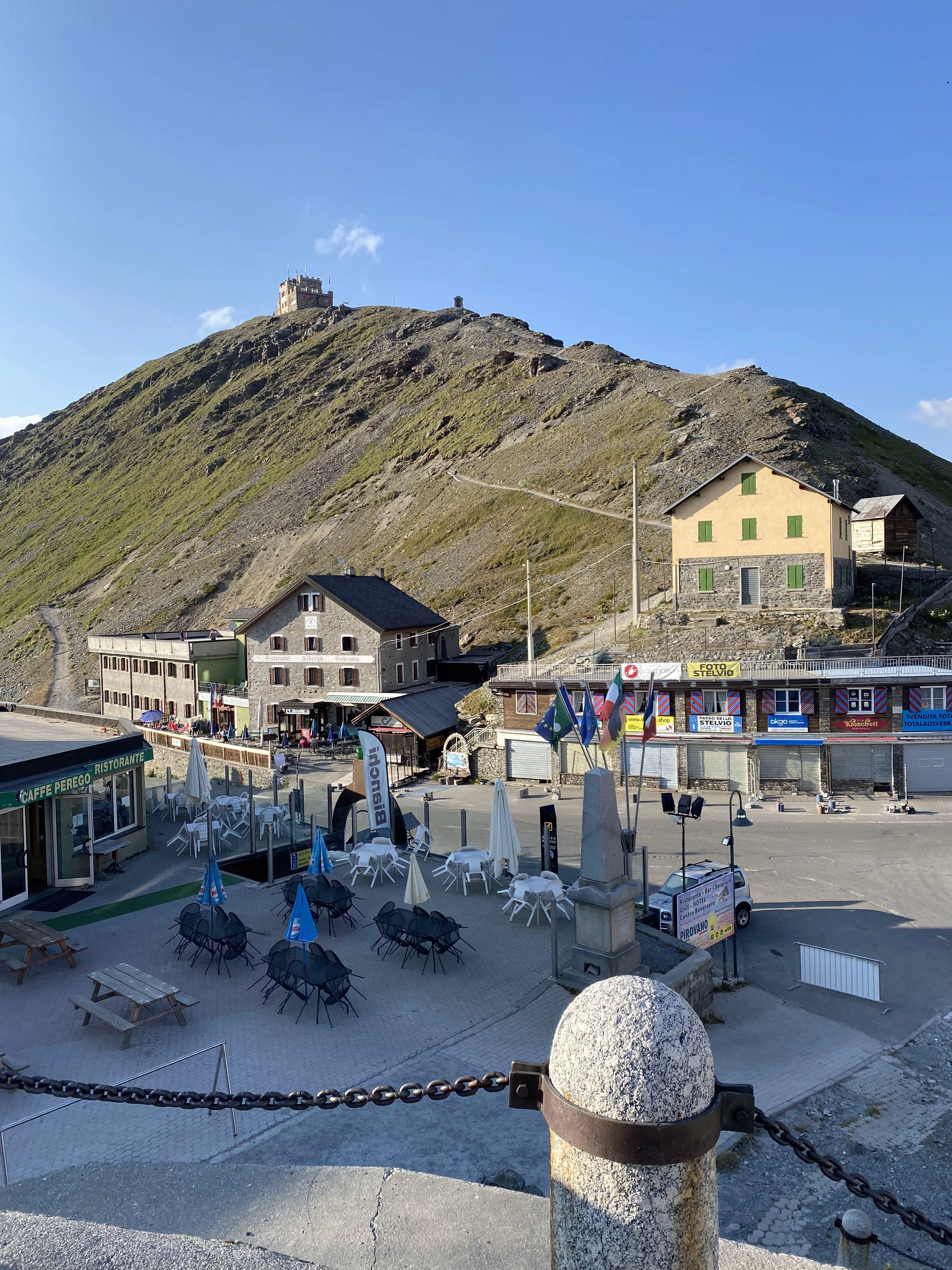 View from the summit of the Stelvio Pass with an empty mountain road and distant snowy peaks under a clear sky.
