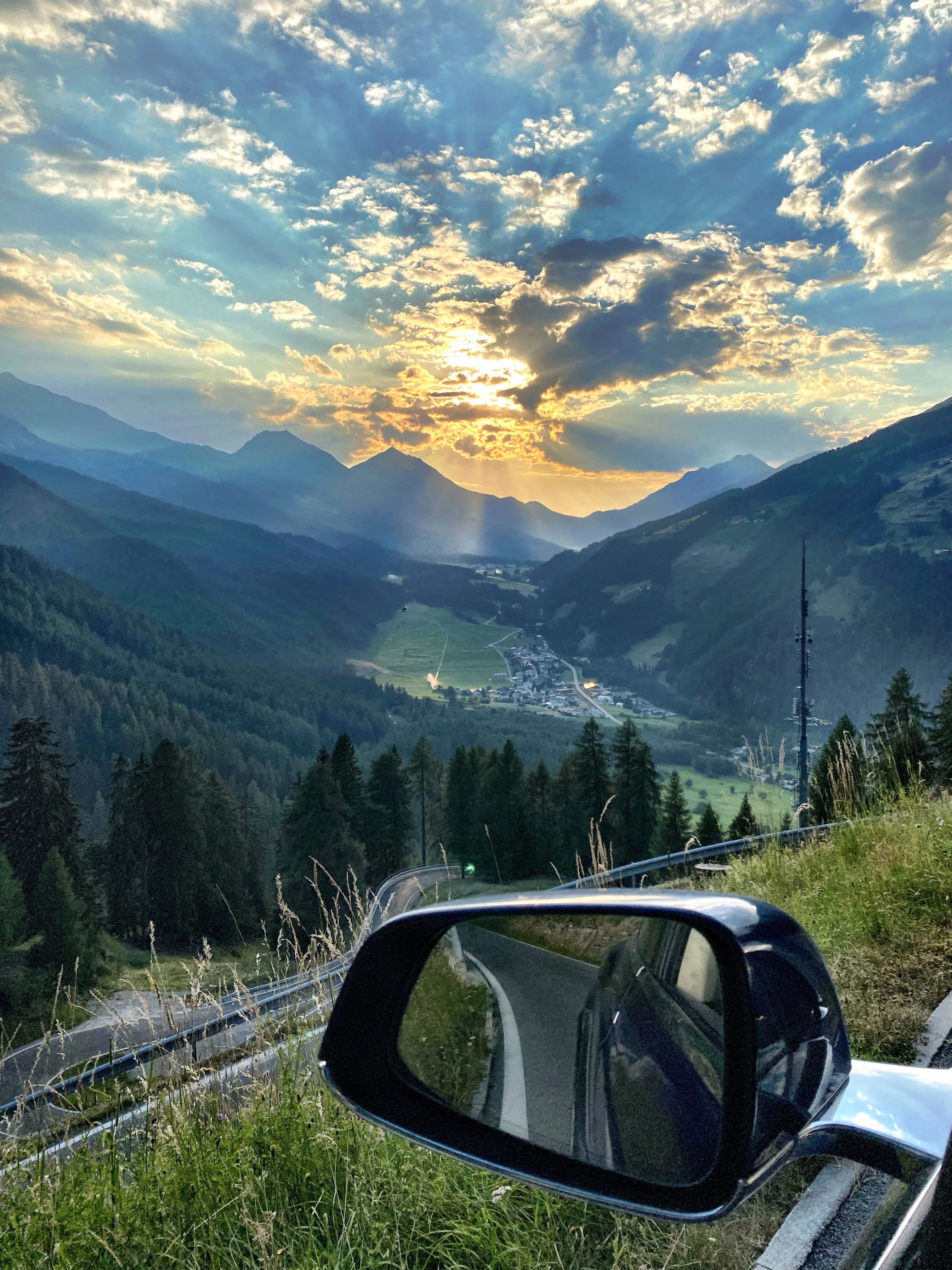 View from a car side mirror showing the Val Müstair valley in golden evening sunlight, with sun rays breaking through the clouds.
